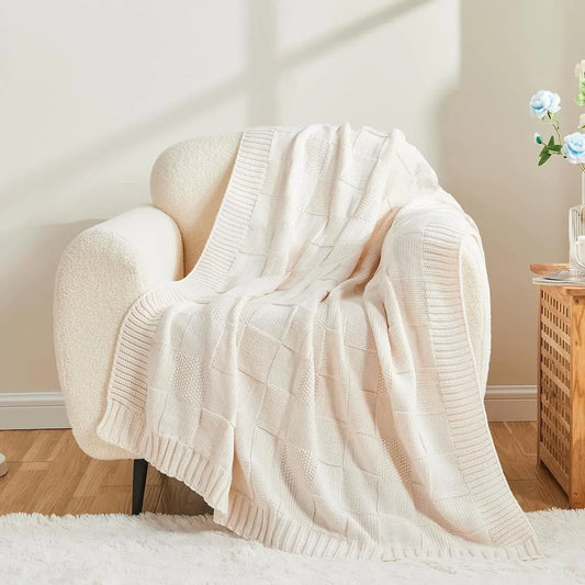 Chunky patchwork knit throw blanket in Ivory Cream draped over a white bouclé armchair in a soft neutral living room with wooden side table