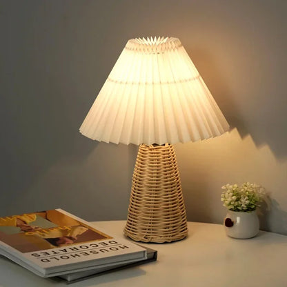 : Rattan & Pleat table lamp with woven rattan base and layered off-white pleated shade, glowing warm light on a desk beside books and a small white ceramic vase with white flowers against a grey wall



