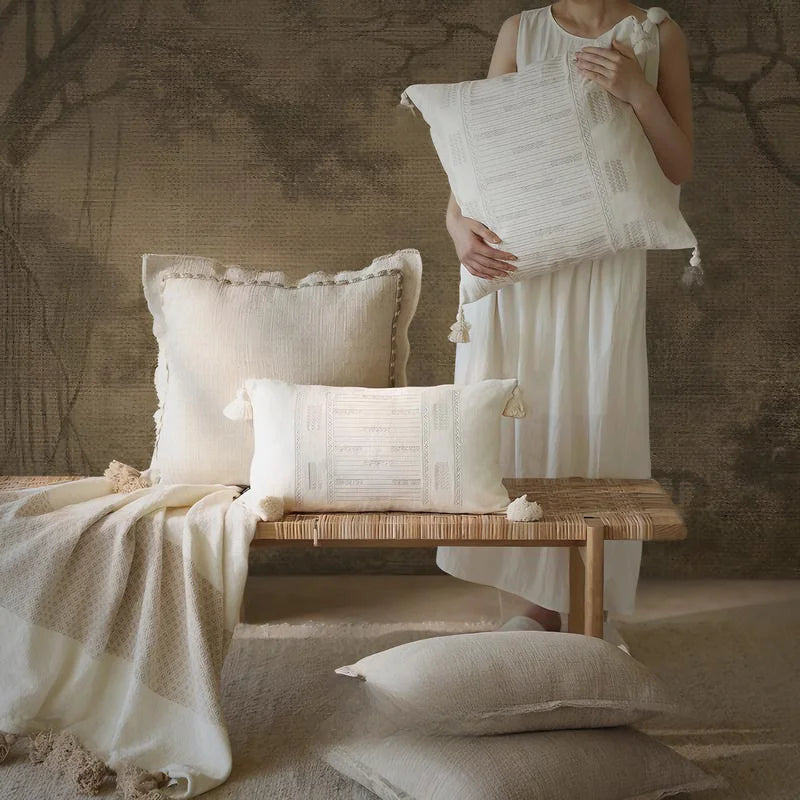 Woman in white linen dress holding an ivory tassel jacquard square cushion cover, styled with a lumbar tassel cushion and natural linen cushion covers on a woven rattan bench, with a linen throw and vintage botanical mural wall — wabi-sabi Japandi home decor.