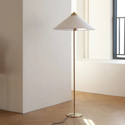 "Cone & Brass" floor lamp with linen shade and brass stem standing on a wooden floor beside a white wall and window