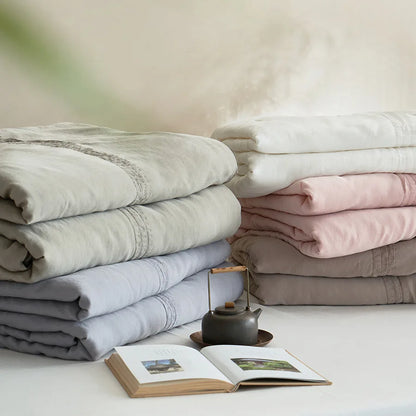 Collection of quilted linen cotton summer throw blankets in ivory white, slate grey, blush pink and warm coffee, neatly stacked with a cast iron teapot and open book in a japandi-style setting