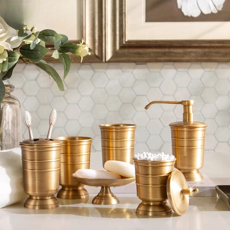 Full antique brass bathroom accessory set styled on white marble counter, including toothbrush holder, tumbler, soap dish, storage jar and soap dispenser, hexagonal tile background with gold mirror and eucalyptus flowers