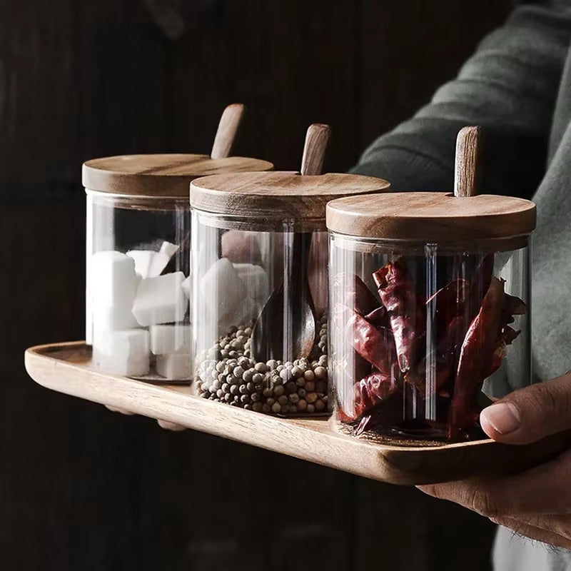  Hands holding an acacia wood tray with three glass spice jars filled with sugar cubes, white peppercorns and dried chillies, wooden spoons resting on each lid, dark moody kitchen background — lifestyle shot of the Acacia & Glass Spice Jar Set by CL House