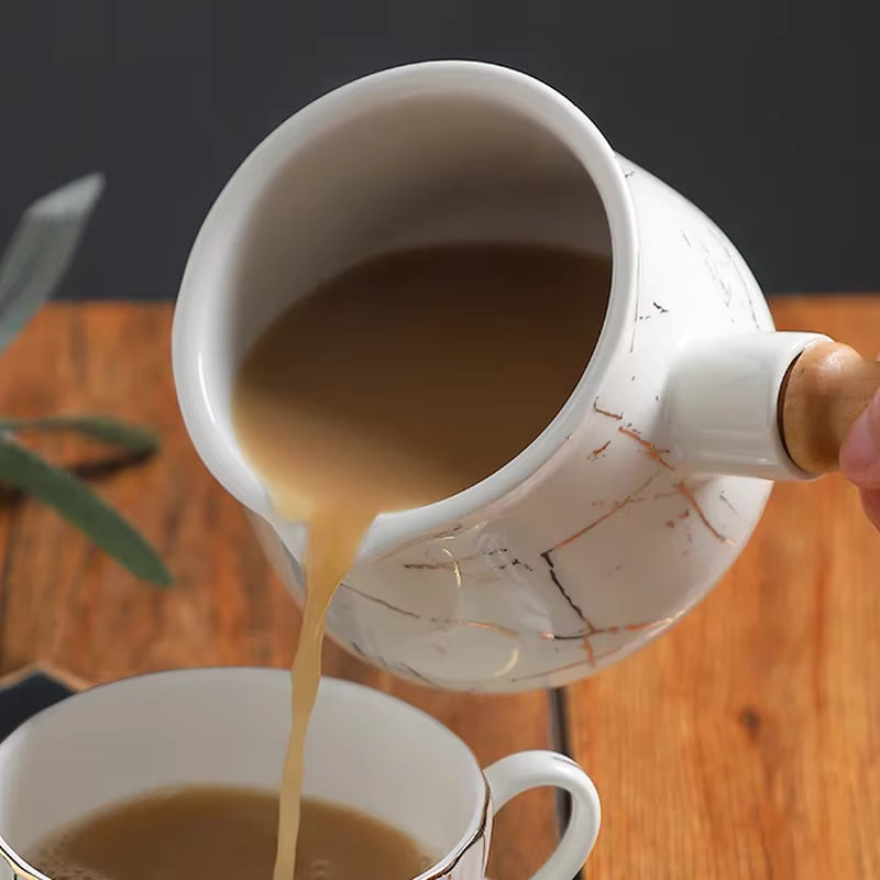  Close-up action shot of white marble ceramic milk jug with gold veining being tilted to pour warm coffee with milk into a white cup, wooden handle visible, rustic wooden surface with olive leaves in the background — lifestyle detail of the Marble Pour Ceramic Milk Jug by CL House