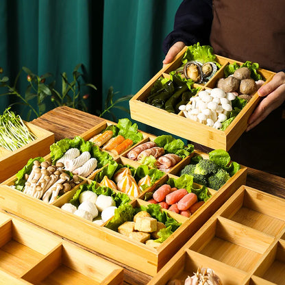  Bamboo Grid trays filled with fresh hot pot ingredients — vegetables, mushrooms, seafood and dumplings — arranged by section, being carried to a wooden table for sharing, lifestyle setting with green backdrop