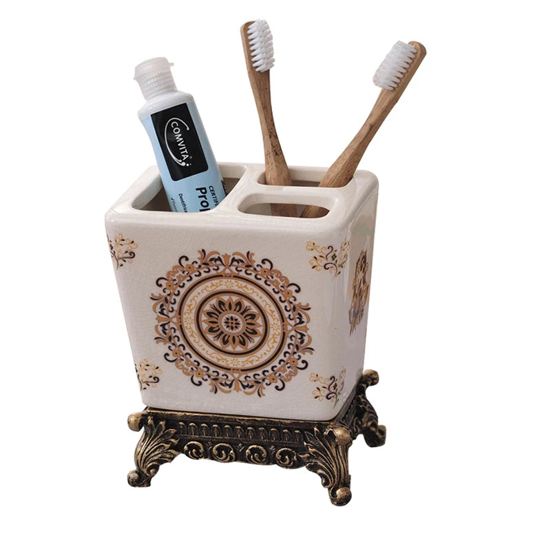 White ceramic toothbrush holder with golden medallion and arabesque pattern, ornate bronze metal base, two bamboo toothbrushes and toothpaste displayed, lifestyle background