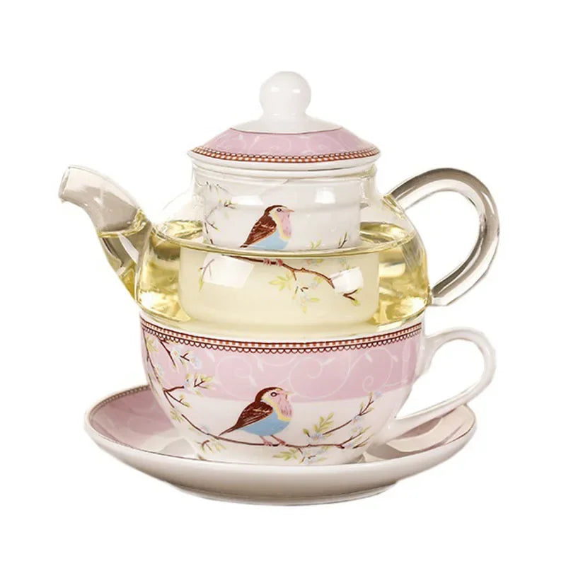  "Happy Bird" tea for one set — glass teapot with porcelain infuser cup and saucer, bird and blossom pattern on a pink background, filled with golden herbal tea, white background.