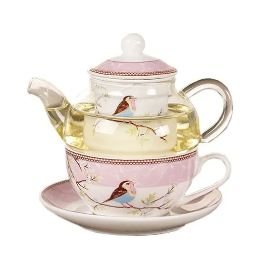  "Happy Bird" tea for one set — glass teapot with porcelain infuser cup and saucer, bird and blossom pattern on a pink background, filled with golden herbal tea, white background.