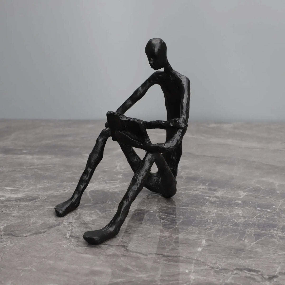 Abstract seated reader sculpture in matte black metal, reading with a book across the knees, on a grey marble surface