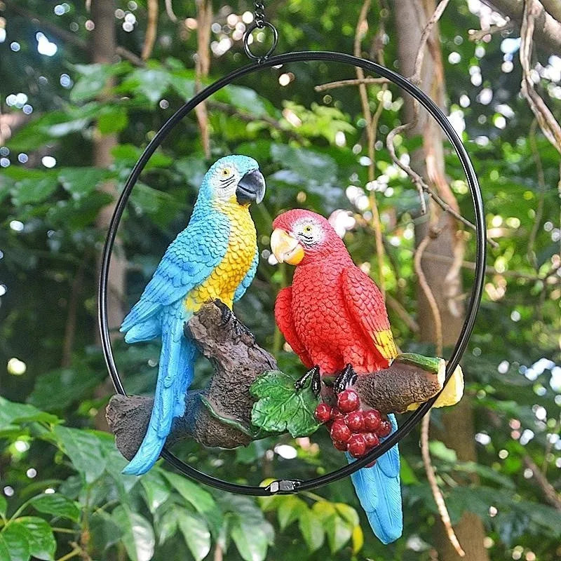 "Perch Duo" resin parrot hanging ring ornament in Scarlet & Azure — full view suspended from tree branch with bokeh garden background, showing hanging hook detail

