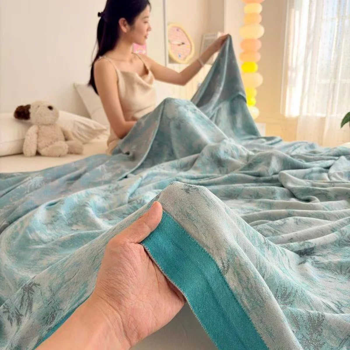  A woman lifting the Teal Garden bamboo fibre jacquard blanket on a bed while a hand in the foreground holds up the soft teal border trim, showcasing the lightweight drape and silky jacquard texture