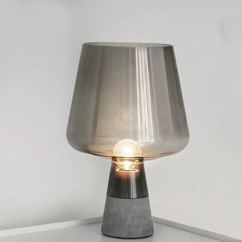 Smoked grey table lamp with a translucent tapered shade and dark textured base, illuminated against a white background.