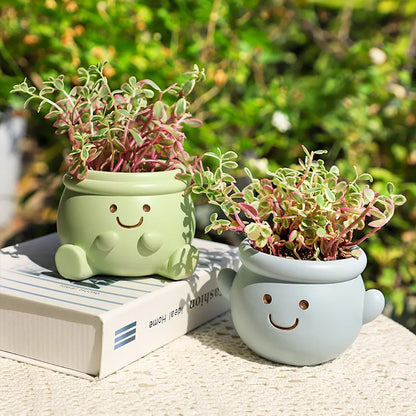 "Happy Pot" smiley face resin succulent planters in sage green and sky blue displayed on fashion and home decor books with lush garden background in warm sunlight