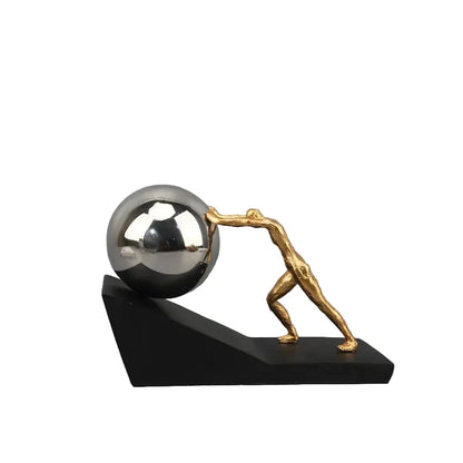 Gold metal figure pushing a reflective silver sphere up a black angled base, shown as a modern Sisyphus sculpture against a grey background.