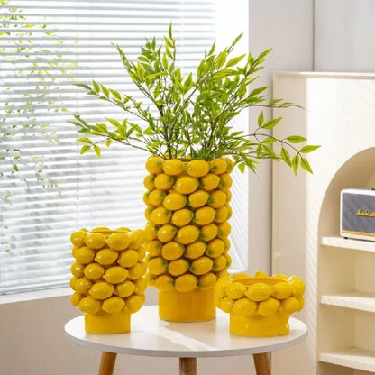 Three yellow ceramic lemon relief vases displayed together on a white round table, large tall vase styled with green bamboo branches, medium and small bowl vases as decorative accents, bright living room with window blinds and white shelving