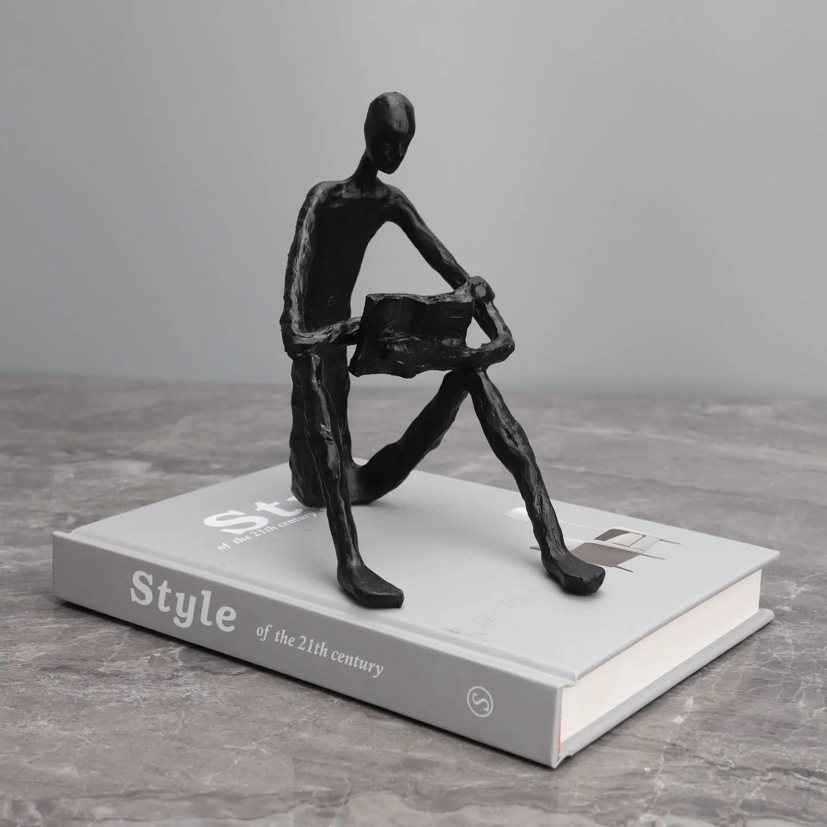 Abstract seated reader sculpture in matte black metal, displayed on a white hardback book on a grey surface