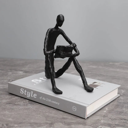 Abstract seated reader sculpture in matte black metal, displayed on a white hardback book on a grey surface