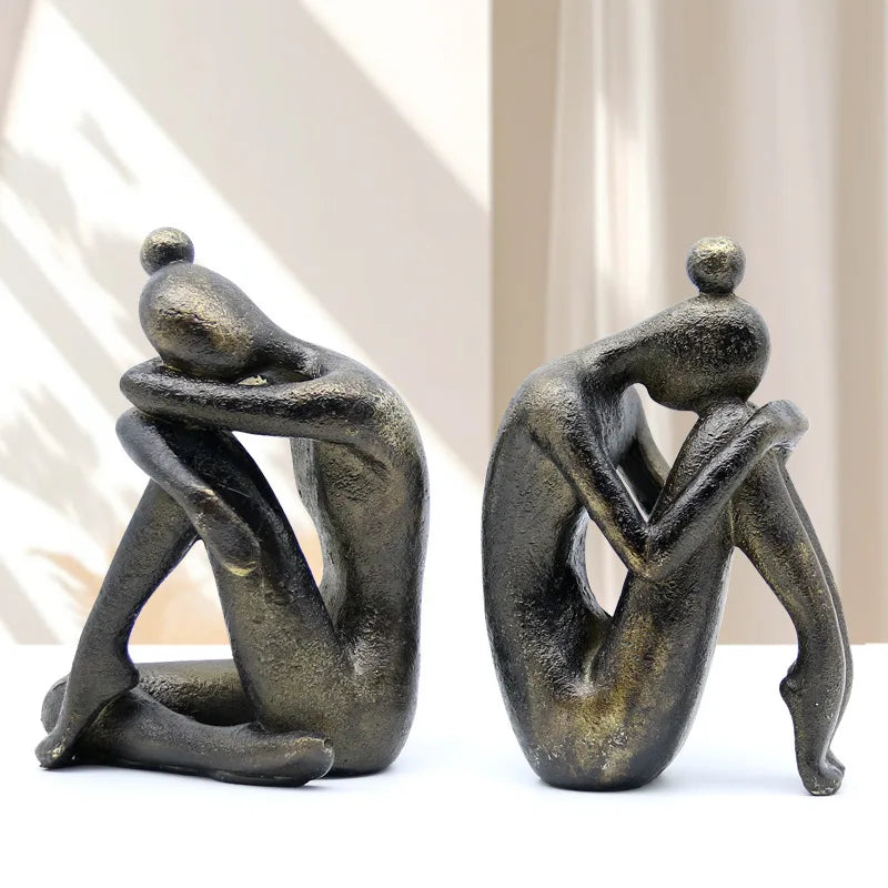 Pair of dark resin abstract seated figures displayed indoors in soft natural light, showing their different sculptural poses and textured finish.