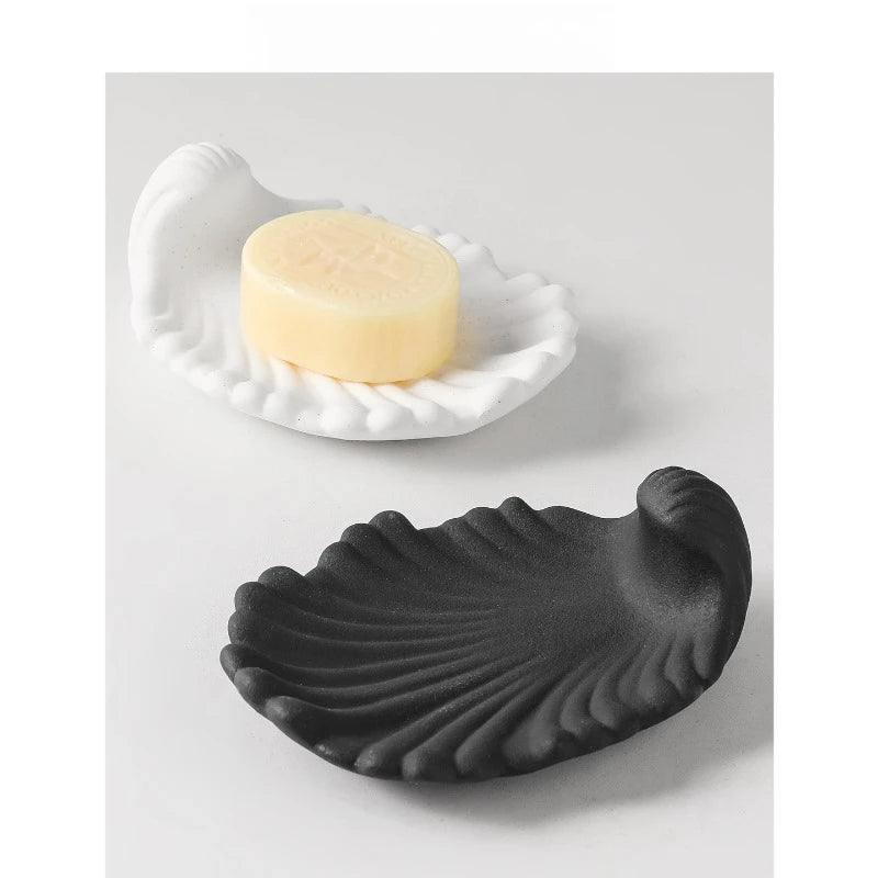 Midnight black and chalk white matte ceramic shell soap dishes displayed together on grey surface, black dish empty in background, white dish holding a bar of soap in foreground