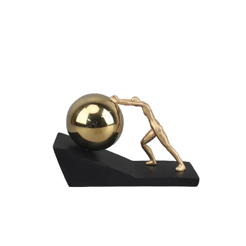 Gold metal figure pushing a reflective silver sphere up a black angled base, shown as a modern Sisyphus sculpture against a grey background.