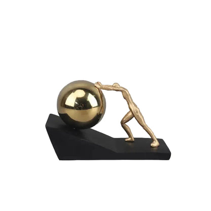 Gold metal figure pushing a reflective silver sphere up a black angled base, shown as a modern Sisyphus sculpture against a grey background.