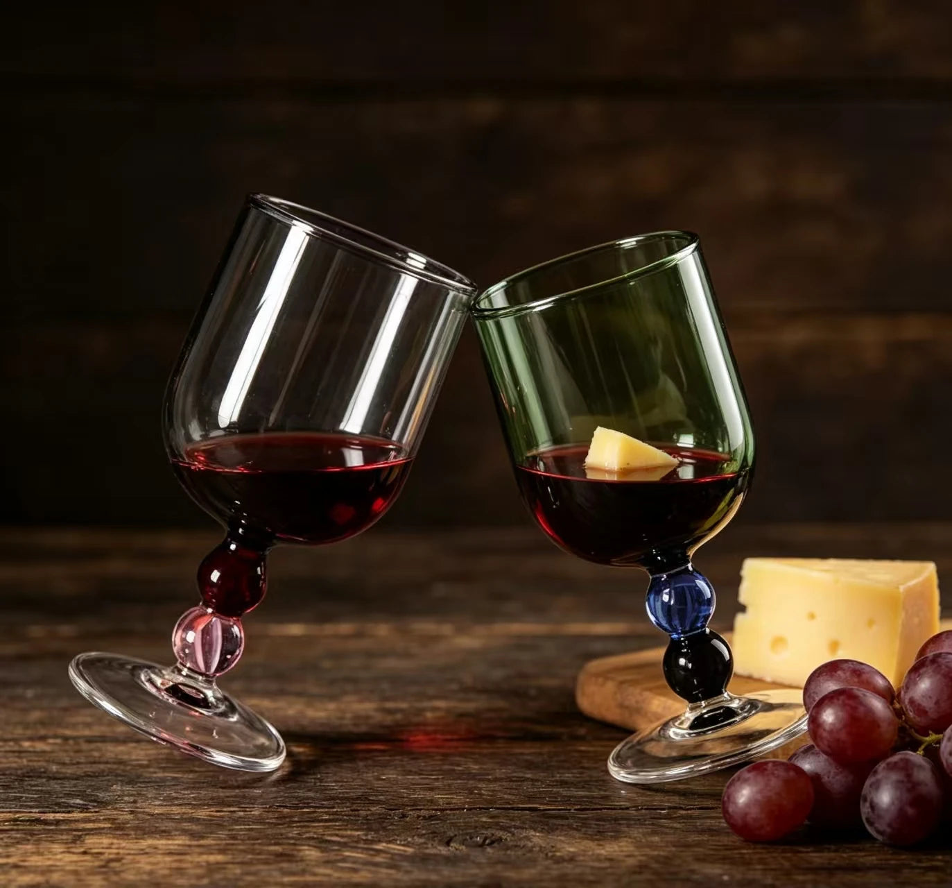 "Chromatic Orb" glass goblets in smoke grey and forest green toasting with red wine, on a rustic wooden table with grapes and cheese