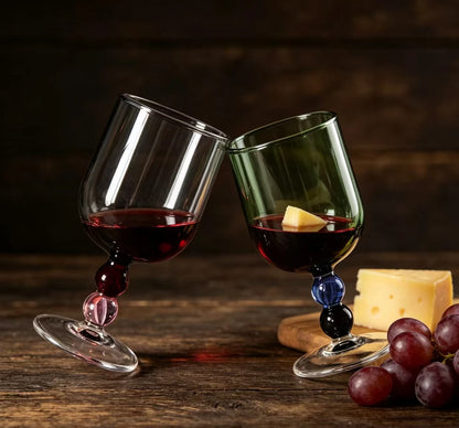 "Chromatic Orb" glass goblets in smoke grey and forest green toasting with red wine, on a rustic wooden table with grapes and cheese