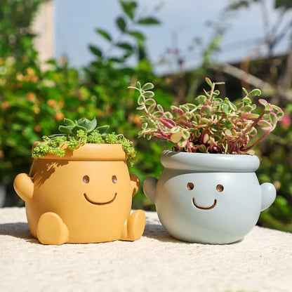 "Happy Pot" smiley face resin succulent planters in mustard yellow and sky blue displayed outdoors on stone surface with lush green garden background