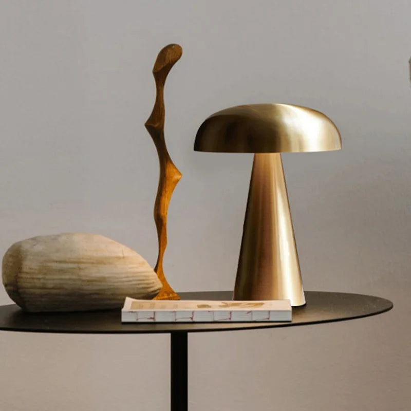 Cap & Glow mushroom table lamp in Golden finish styled on a round black side table alongside a wooden sculptural figurine and a smooth stone object, creating a curated, editorial home decor vignette.