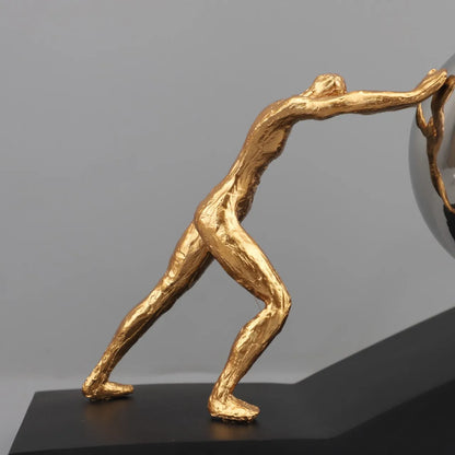 Close-up of the gold metal figure leaning forward to push a reflective sphere on The Ascent black sculptural base.