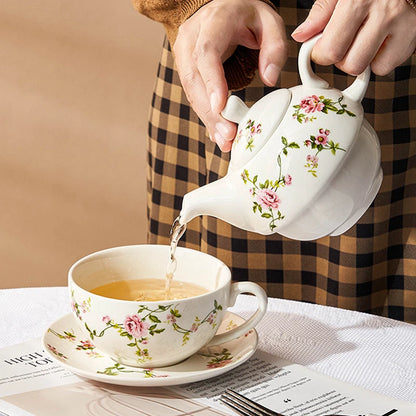 Hands pouring tea from a white porcelain teapot with pink rose and green vine pattern into a matching cup and saucer on a white table, warm cosy atmosphere