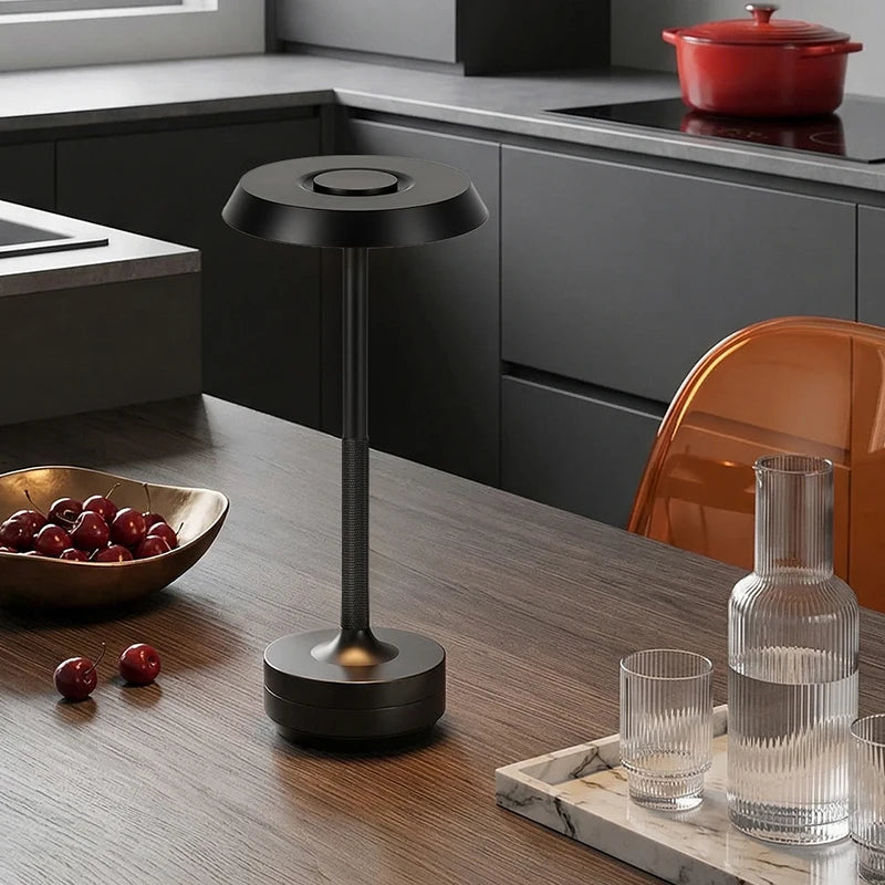 Black rechargeable table lamp on a dark wood dining table in a modern kitchen, styled with a bowl of cherries and glass carafe — "Disc & Stem" by CL House