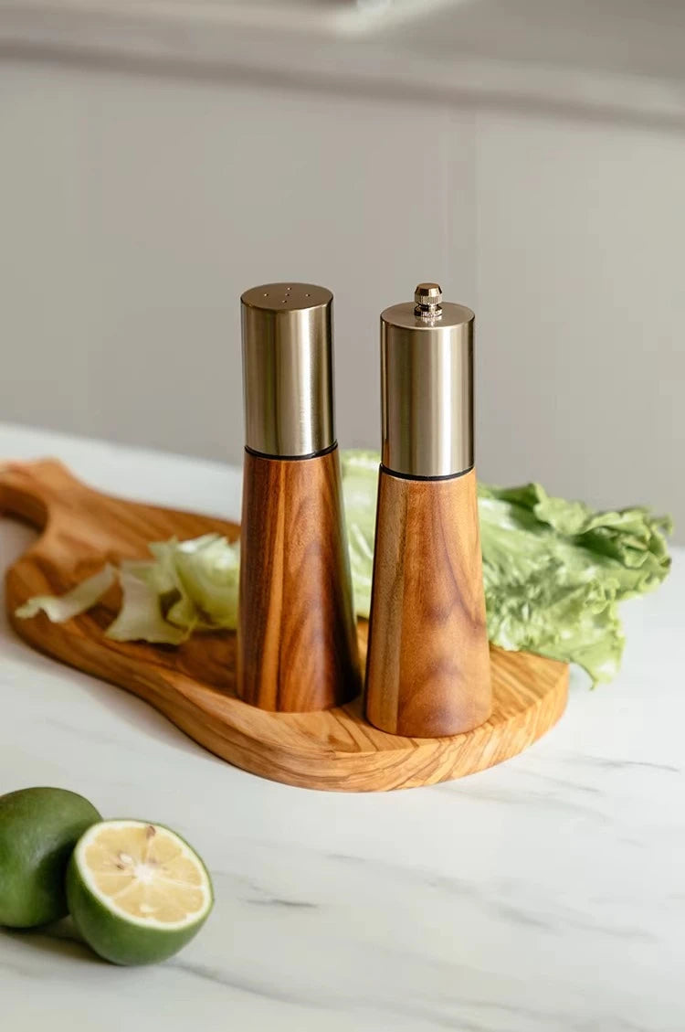 Acacia Twist salt and pepper mill duo styled on a wooden chopping board with fresh lettuce and limes on a white marble kitchen counter