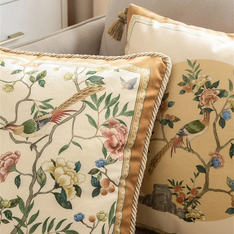 : Close-up detail of two "Golden Garden" chinoiserie velvet cushion covers styled together, exotic birds and peonies on ivory ground with braided gold rope border — CL House Cushions collection
