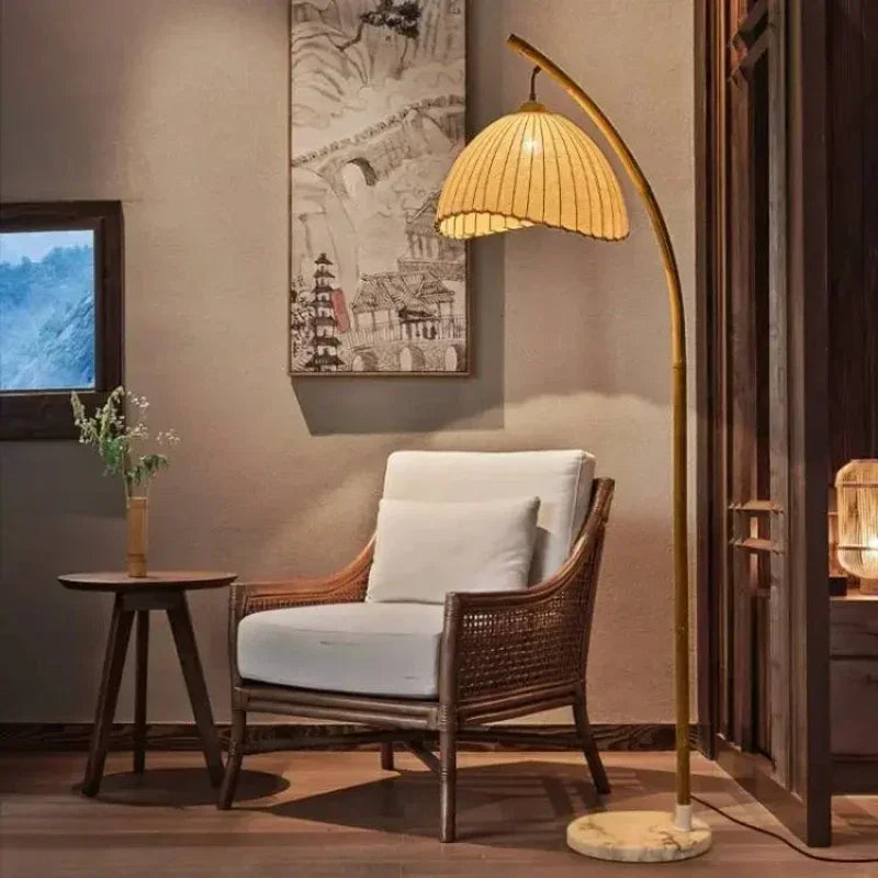 Imagem 4: Arc & Pleat canvas floor lamp with marble base arching over a rattan armchair with white cushion, in a warm japandi-style living room with Chinese ink painting on the wall