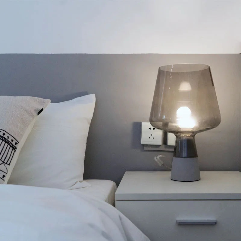  "Smoke & Cement" glass table lamp with smoke grey shade glowing on a white bedside table in a minimal bedroom setting