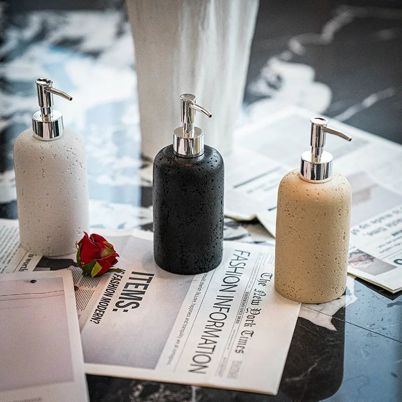 : Three Travertine Curve soap dispensers — Chalk Stone, Midnight Stone, and Rose Stone — styled on dark marble surface with fashion magazines and red rose, chrome pumps catching the light
