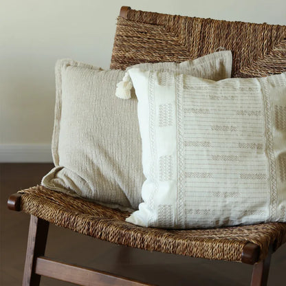 Ivory tassel jacquard lumbar cushion cover with geometric embroidery and hand-knotted corner tassels, styled with a natural linen square cushion cover on a woven seagrass bench in a minimal neutral interior.