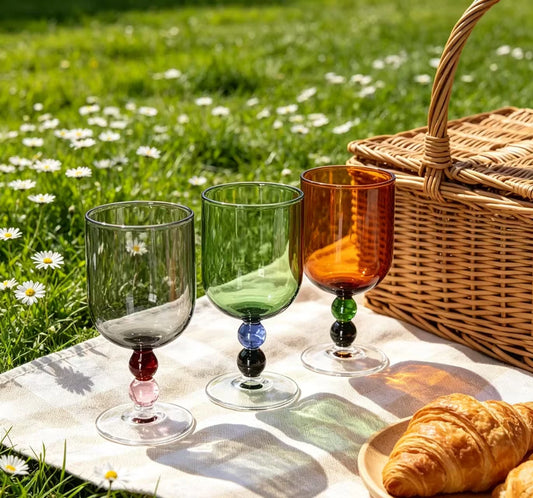 "Chromatic Orb" glass goblets in smoke grey, forest green and amber, styled on a picnic blanket with a wicker basket and croissants
