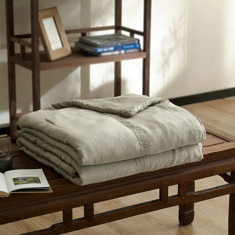 Sage green quilted linen cotton summer throw blanket, neatly folded on a dark wood bench in a japandi-style interior with wabi-sabi books on shelving and ceramic vase