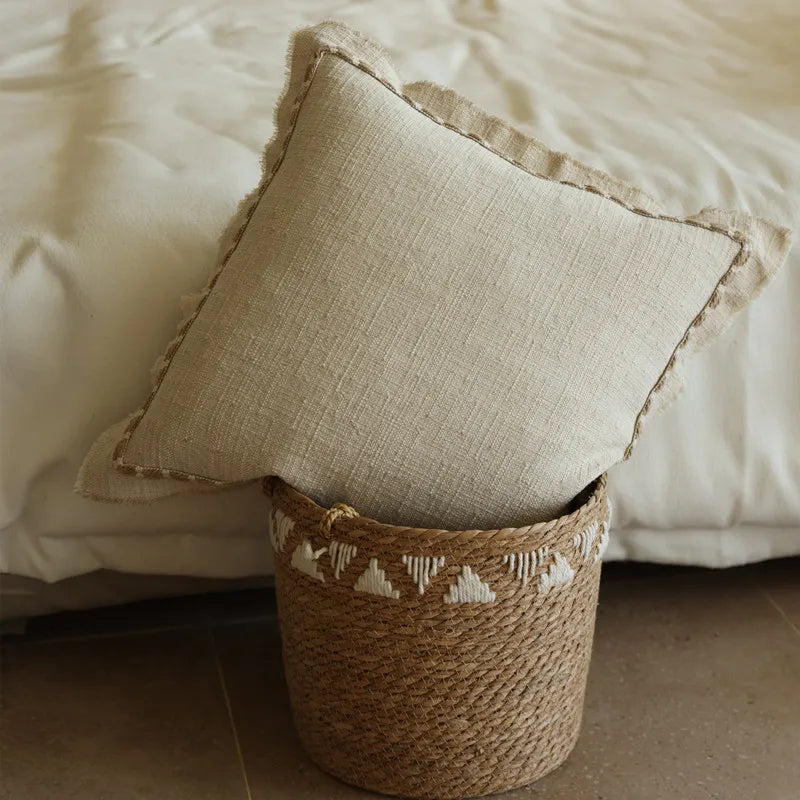 Natural linen cushion cover with delicate fringe edge, leaning against a white bed, resting on a handwoven seagrass basket, styled in a calm and earthy wabi-sabi bedroom setting.
