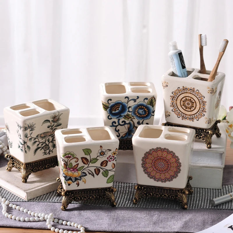  Five floral ceramic toothbrush holders with ornate bronze metal bases displayed together, showing Garden Bird, Peony Garden, Blue Chinoiserie, Crimson Bloom and Golden Medallion designs, white background with pearl beads