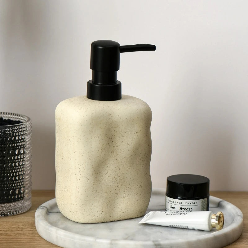 Sand Form ceramic soap dispenser with matte black pump on marble tray, styled with Sea Breeze fragrance candle and skincare tube, glass candle holder on left, warm neutral background