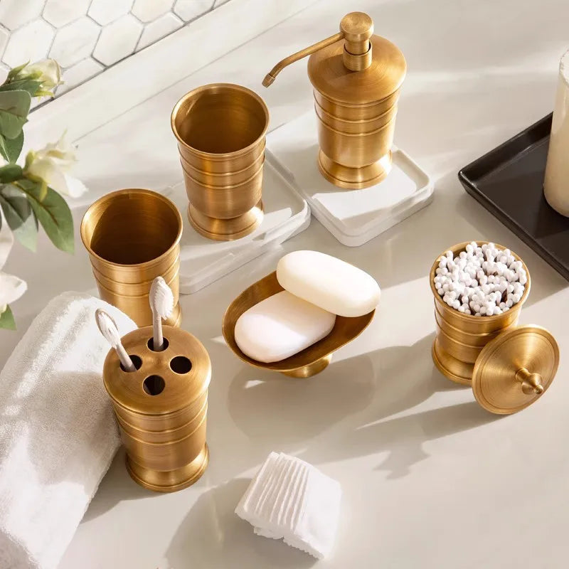 Antique brass bathroom accessory set arranged on white marble counter in natural light, showing toothbrush holder, tumbler, soap dish with bar soap, storage jar with cotton buds and soap dispenser, white towel and eucalyptus leaves