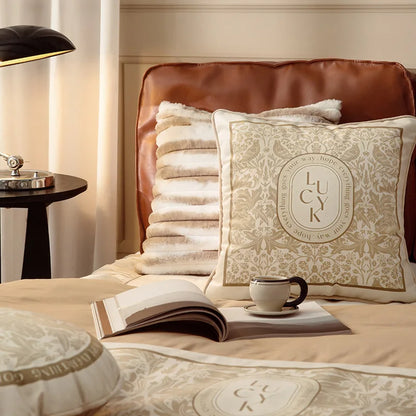 "Lucky Bloom" botanical velvet square cushion cover styled on a leather headboard bed in a warm neutral bedroom, golden floral damask with LUCK medallion, open book and ceramic cup in foreground — CL House Cushions collection