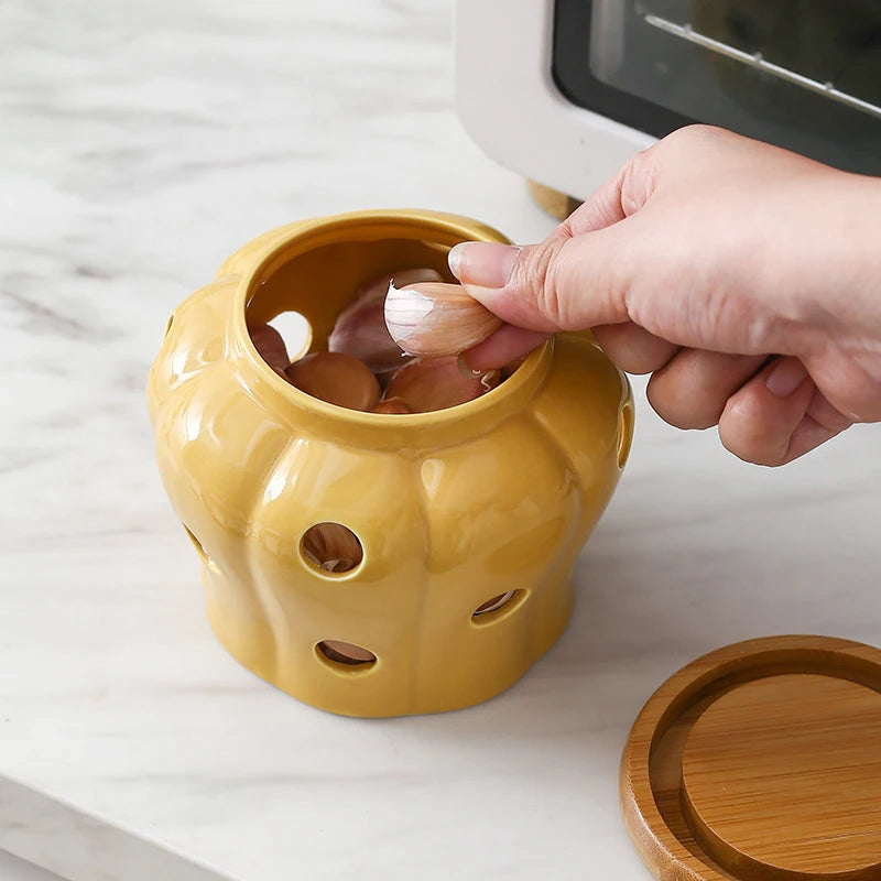 Hand placing a garlic clove into a mustard yellow pumpkin-shaped ceramic storage jar with ventilation holes and bamboo lid beside it, CL House & Home