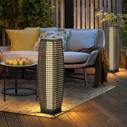 Two dark wicker solar floor lanterns glowing with warm amber light on a wooden deck terrace beside a rattan sofa with yellow and floral cushions at night