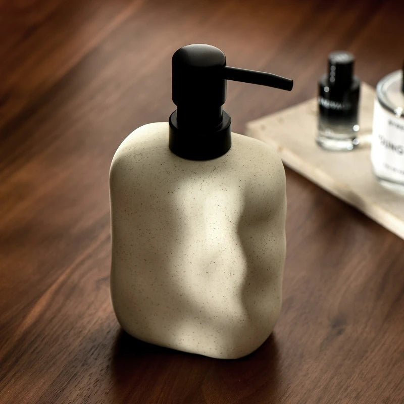  Sand Form ceramic soap dispenser with matte black pump on dark walnut surface, styled with perfume bottle and glass skincare jar on linen tray in background