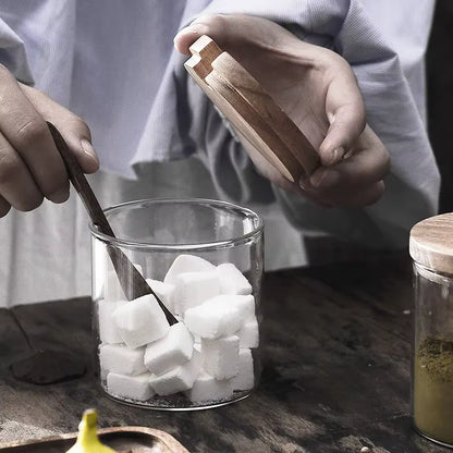 Hands opening an acacia wood lid from a glass jar filled with white sugar cubes, wooden spoon stirring inside, rustic dark wooden countertop with a second spice jar visible in the background — lifestyle detail of the Acacia & Glass Spice Jar Set by CL House