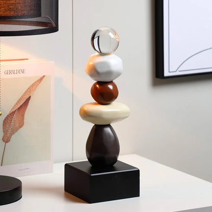 Abstract ceramic stacked sculpture with a clear upper sphere, white and brown rounded forms, a cream oval element and a black square base, displayed on a white sideboard in a modern interior.